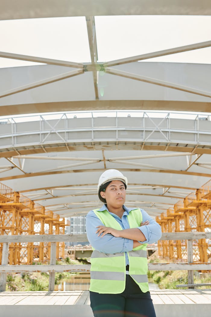 Female engineer with arms crossed at a modern construction site, wearing protective gear.